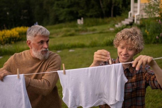 Elderly man and teenage boy joyfully hanging laundry in a sunny garden setting.